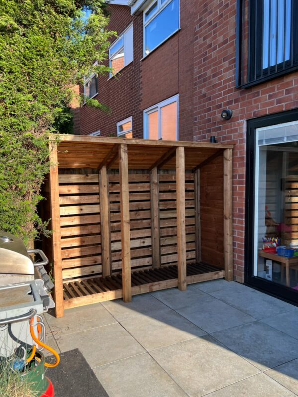 8ft log store Holton with slatted back and forward-incline roof, shown in garden against brick wall.